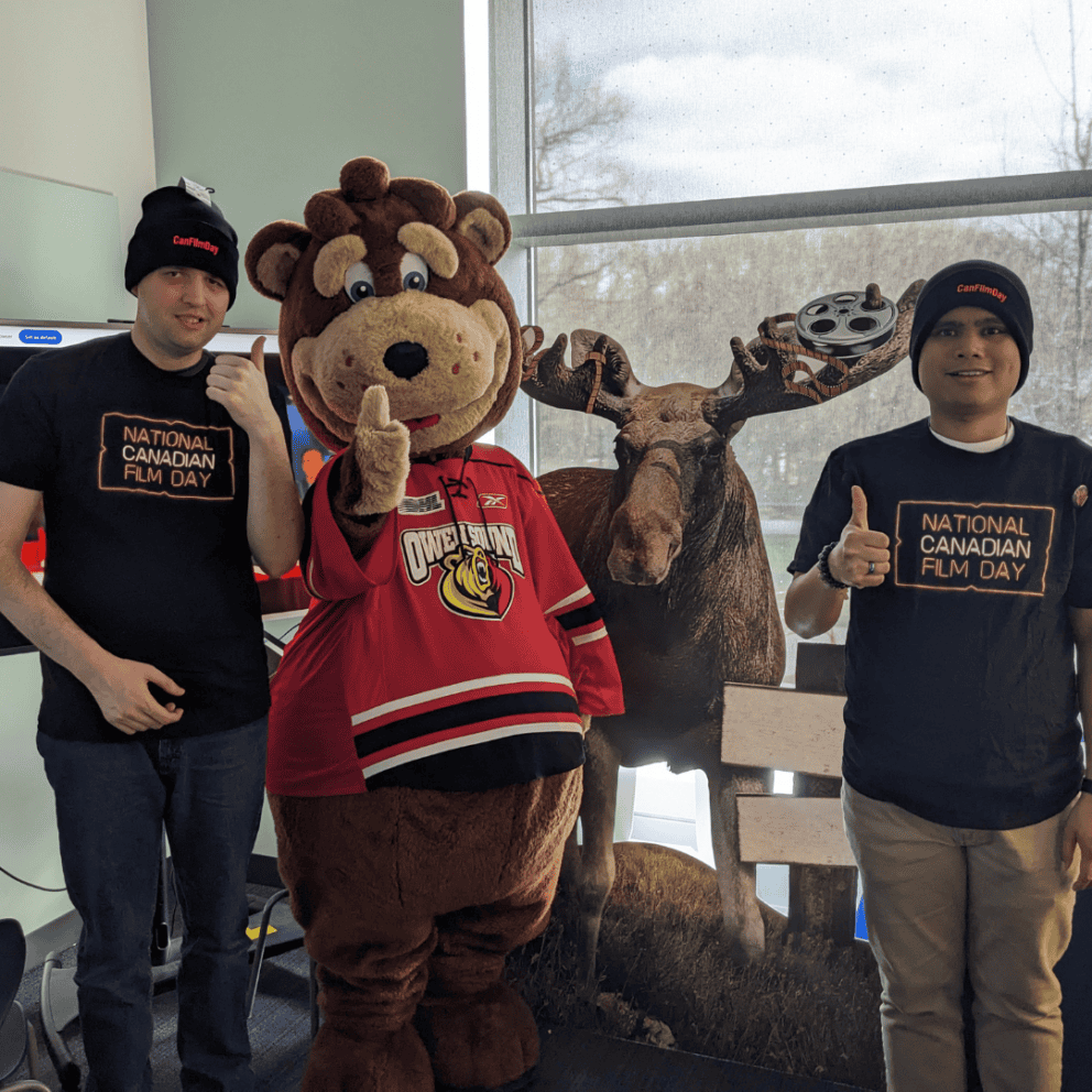 Two young men in CanFilmDay toques stand with a mascot and a cardboard moose.